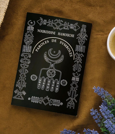 Le livre Paroles De Symboles par Noureddine Hamouche posé sur une table à coté de fleur et tasse de café - sur Azamoul mode et accessoires berbères amazigh.