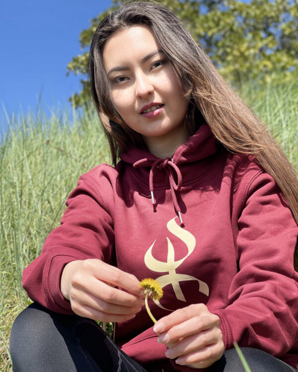 sweat à capuche YAZ avec le symbole berbère amazigh YAZ , symbole du peuple libre amazigh, par azamoul mode et accessoires berbères de couleur bordeaux, portée par une femme aux cheveux longs qui tient une fleur  dans un champ